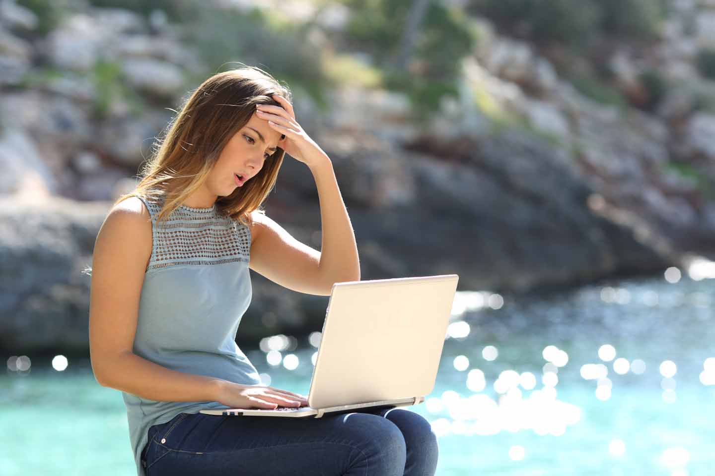 Junge Frau sitzt mit einem Laptop am Meer und schaut mit verärgertem Gesichtsausdruck auf den Bildschirm.