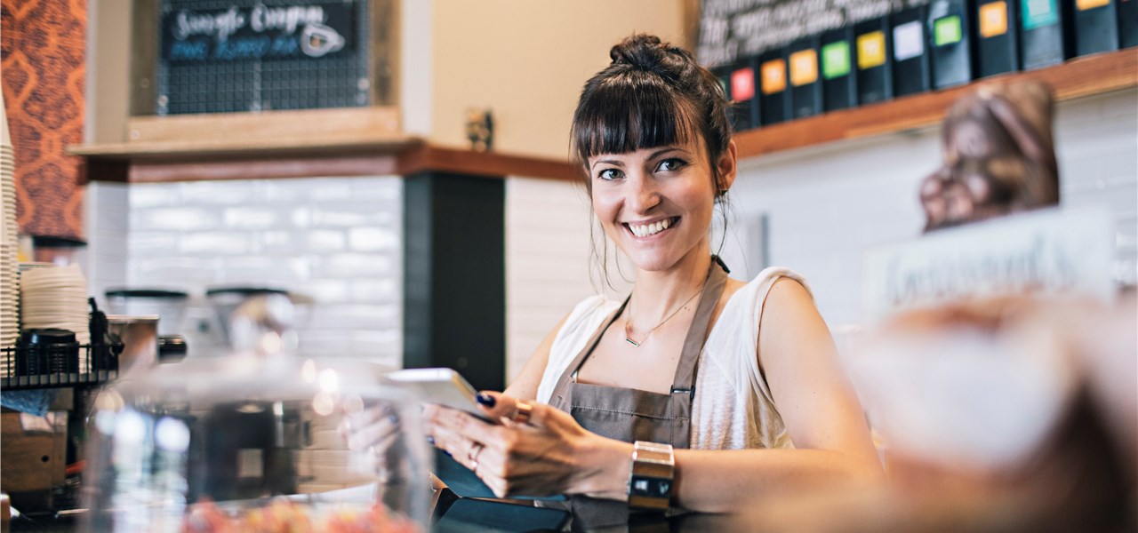 Eine Kaffeebesitzerin mit Schürze lächelt fröhlich in die Kamera. 