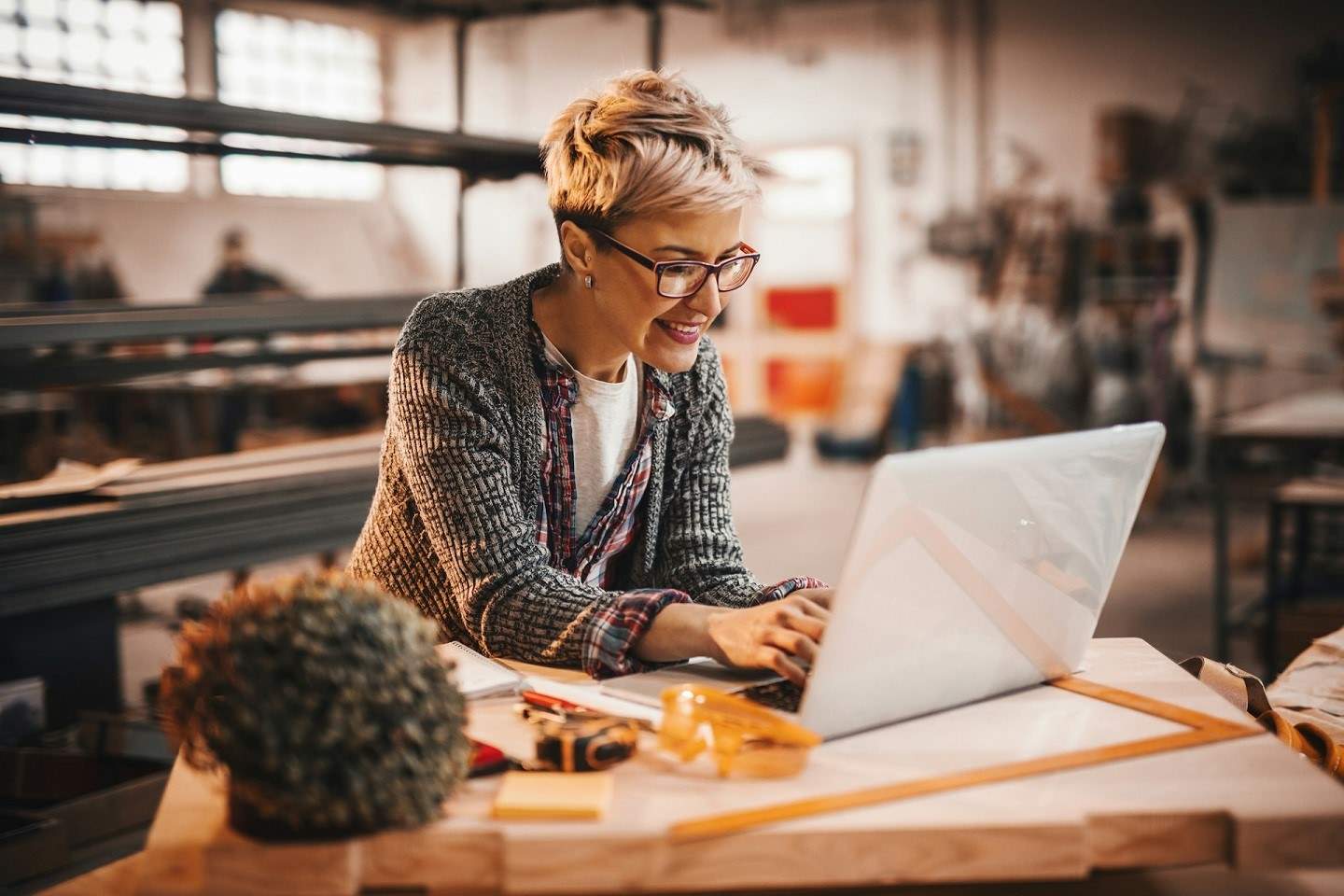 Eine Frau sitzt im Businessoutfit in einer Produktionshalle am Laptop.