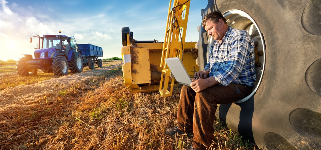 Ein Landwirt sitzt im Reifen seines Treckers und sieht in einen Laptop. 