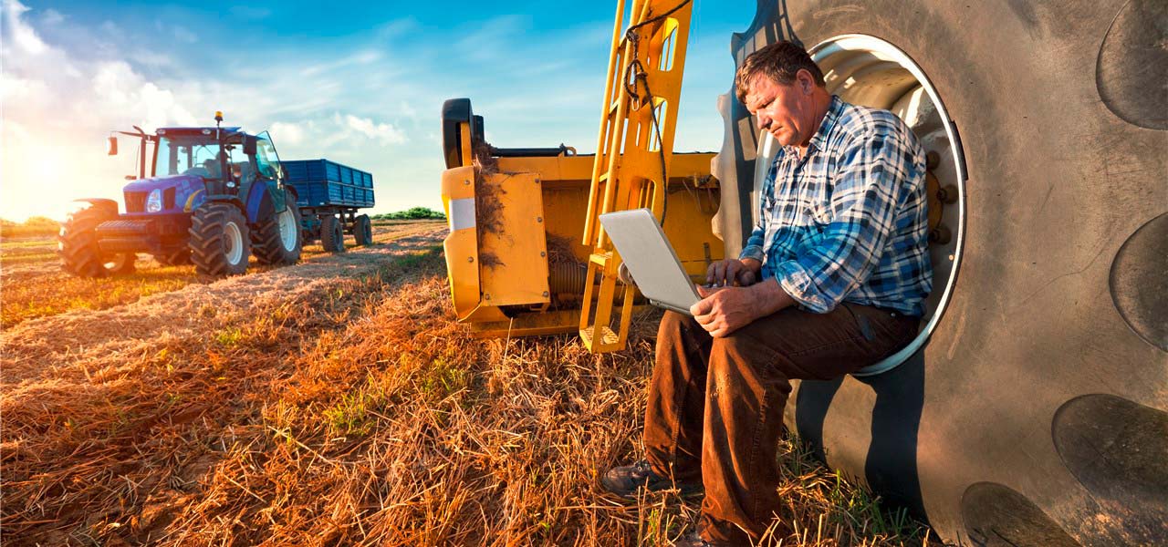 Ein Landwirt sitzt im Reifen seines Treckers und sieht in einen Laptop. 