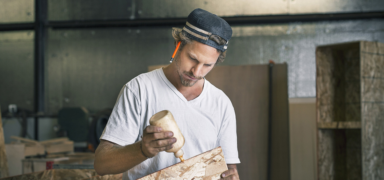 Ein Schreiner trägt konzentriert Leim auf ein Holzbrett in seiner Werkstatt auf.