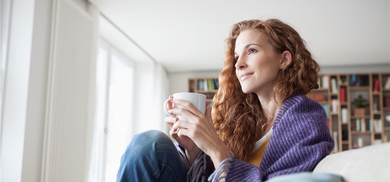 Junge Frau sitzt gemütlich auf dem Sofa mit einer Tasse in der Hand.