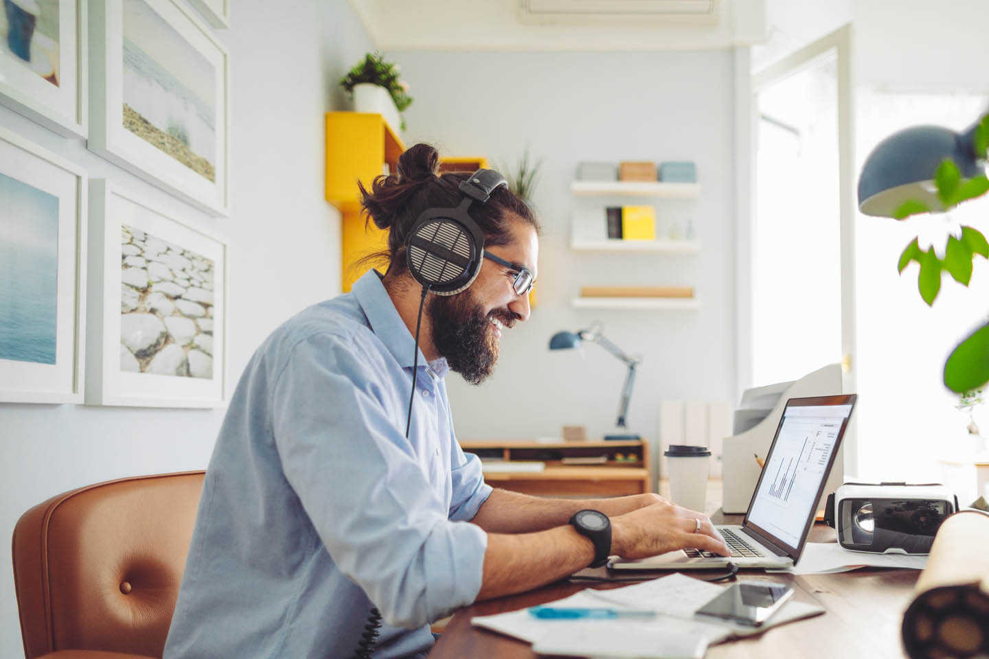 Ein bärtiger Mann mit Headset arbeitet zuhause im Arbeitszimmer am Laptop. 