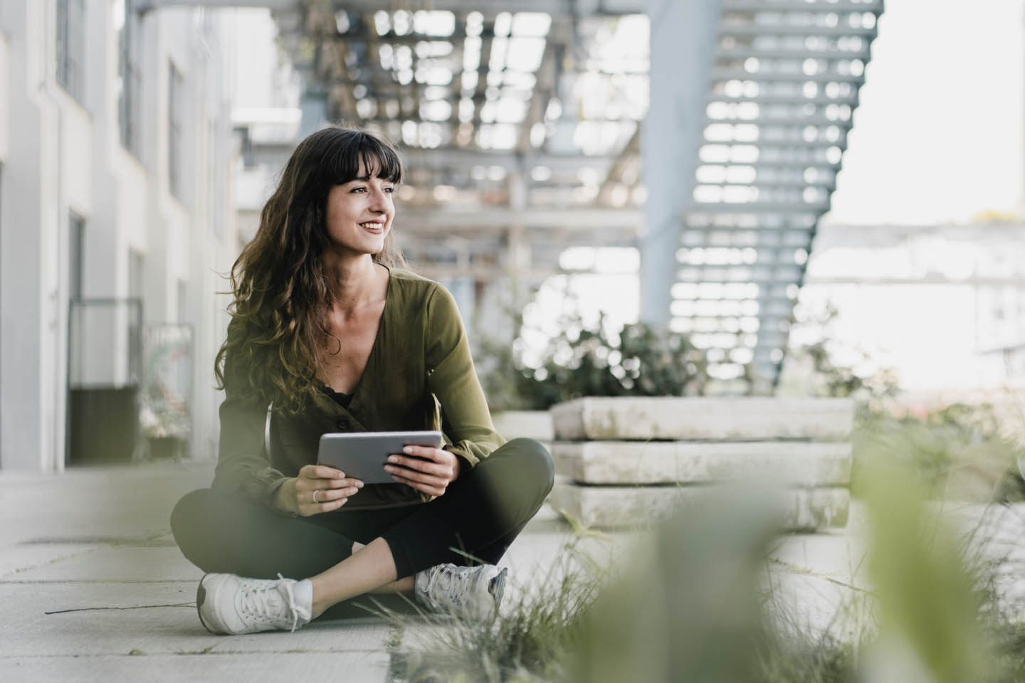 Eine Frau sitzt in einem Industrieviertel mit dem Tablet im Schneidersitz am Betonboden.