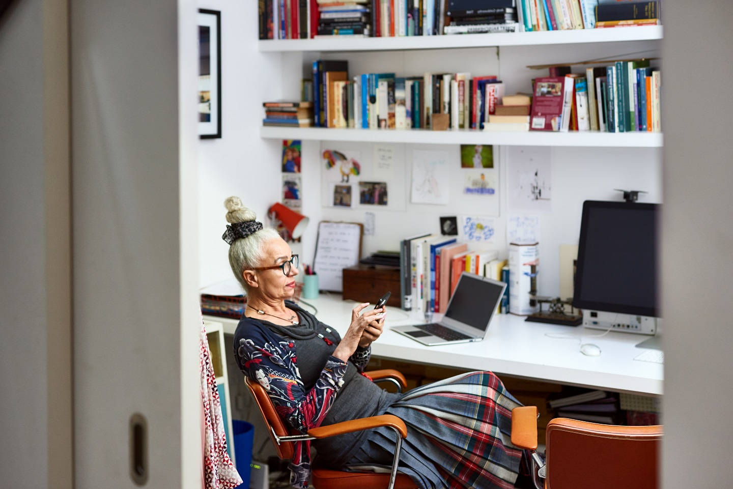 Boho-Frau in den 60ern, die am Schreibtisch mit dem Handy simst, Arbeit von zu Hause, arbeitende Senioren, Kleinunternehmen