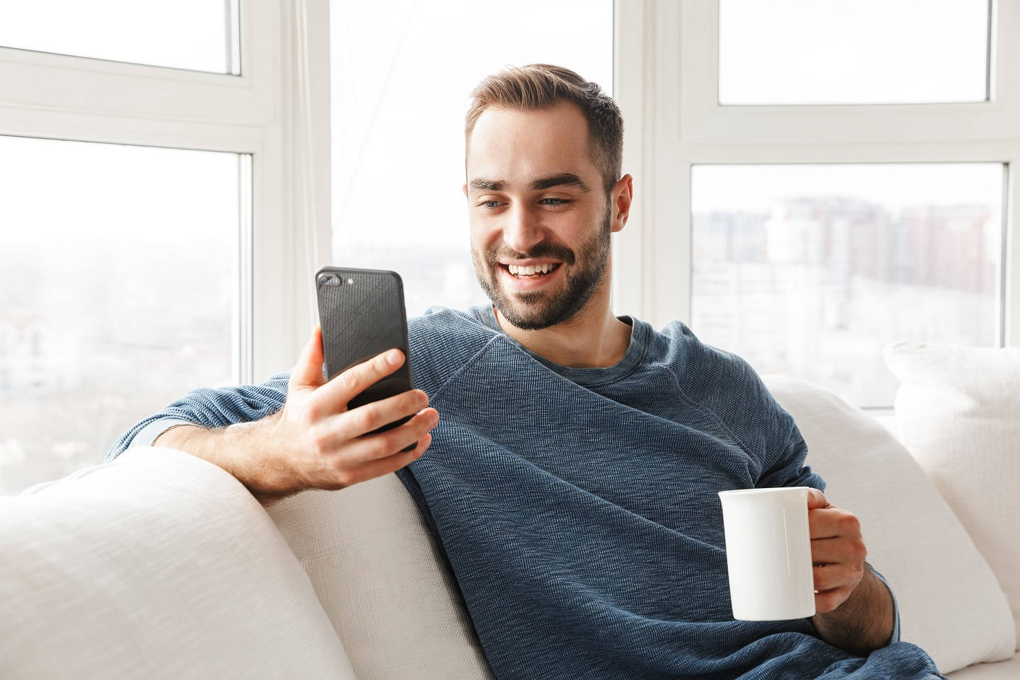 Attraktiver junger Mann entspannt sich zu Hause auf einer Couch, benutzt sein Handy und trinkt Kaffee;