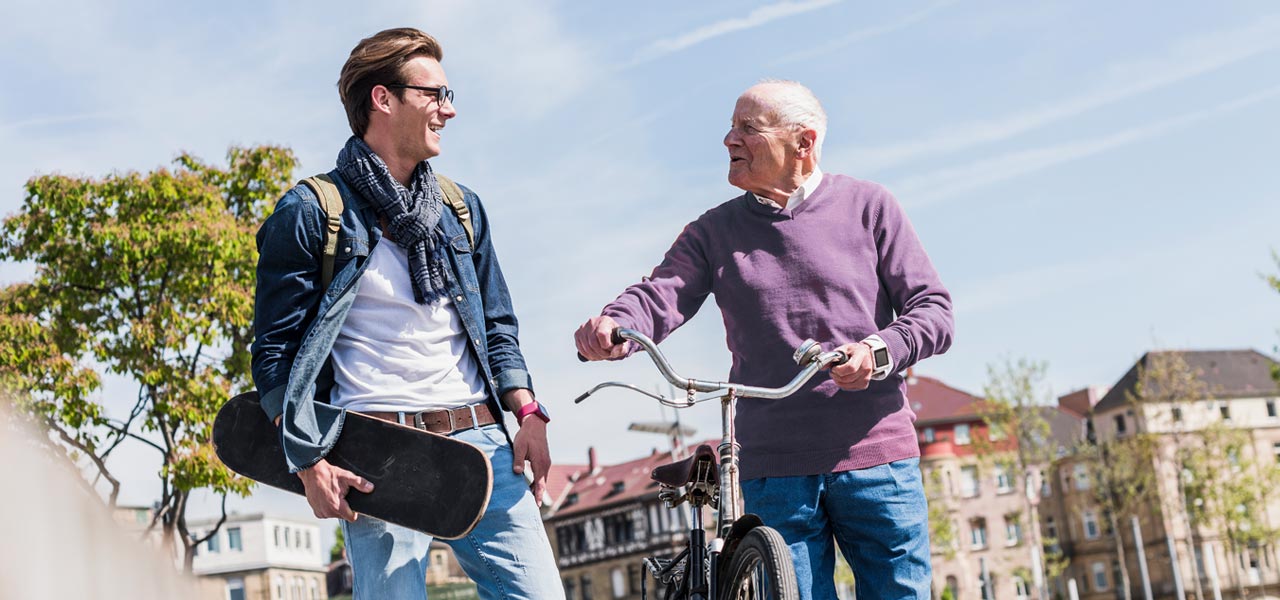 Älterer Mann mit Fahrrad unterhält sich angeregt mit jüngerem Mann, der ein Skateboard trägt.