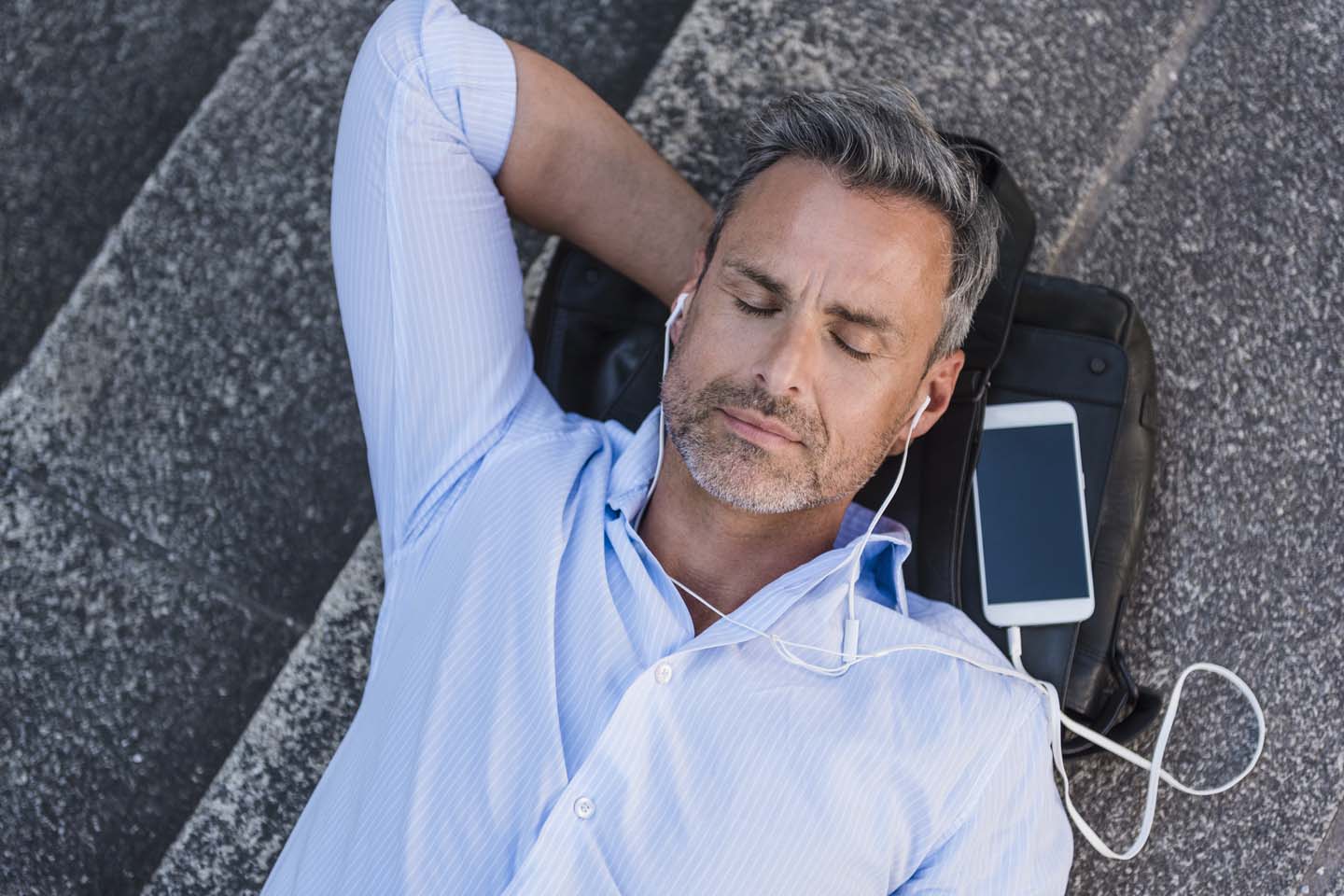 Mann mit geschlossenen Augen liegt auf einer Treppe, mit Handy und Kopfhörern.