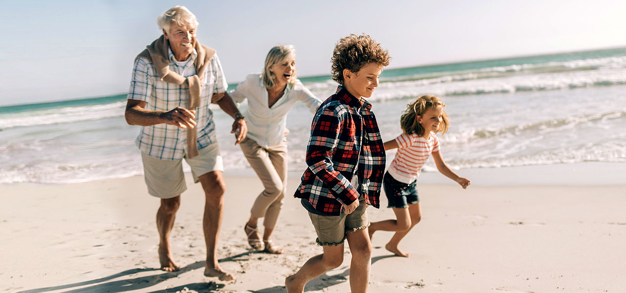 Großeltern rennen lachend mit zwei Kindern am Strand entlang.