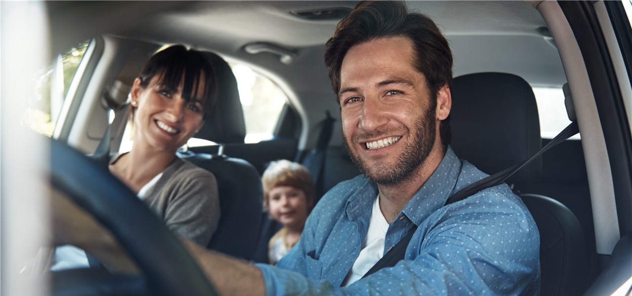 Eine junge Familie im Auto grinst in die Kamera.