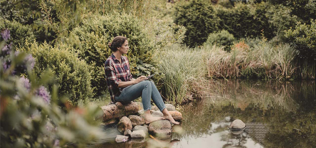 Eine Frau sitzt an einem kleinen Teich und streckt die Zehenspitze ins Wasser.