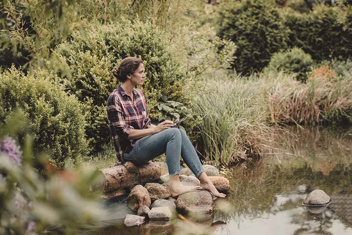 Eine Frau sitzt an einem kleinen Teich und streckt die Zehenspitze ins Wasser.