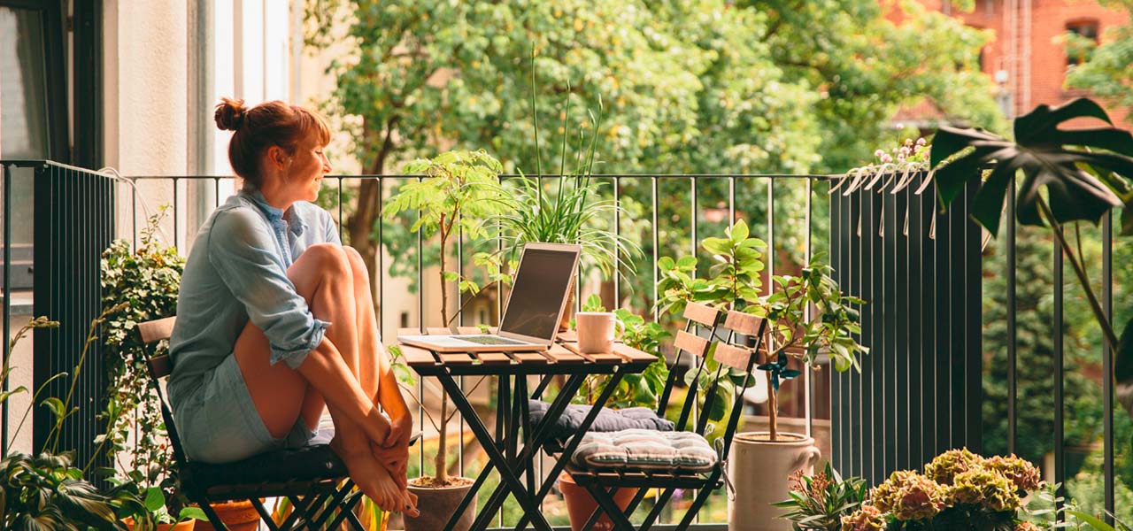 Auf einem Balkon, umgeben von viel Grün sitzt eine Frau mit Laptop. 