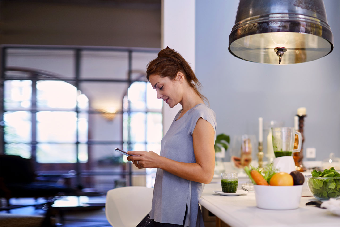 Eine Frau steht in ihrer Küche an die Ablage gelehnt und schaut aufs Handy.