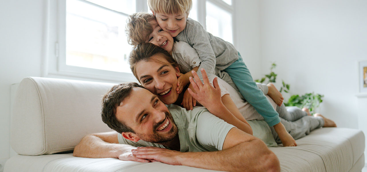 Vater, Mutter und zwei Kinder liegen übereinander auf dem Sofa und lachen.