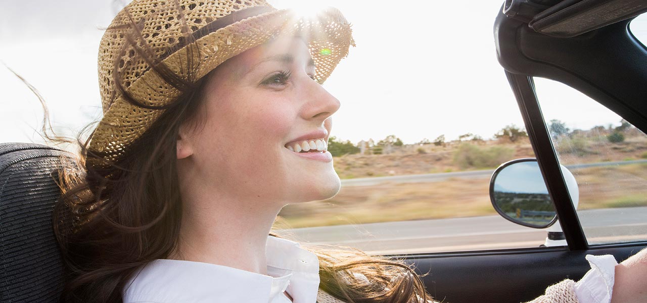 Junge Frau mit Sonnenhut fährt im Cabrio durch eine südliche Landschaft.