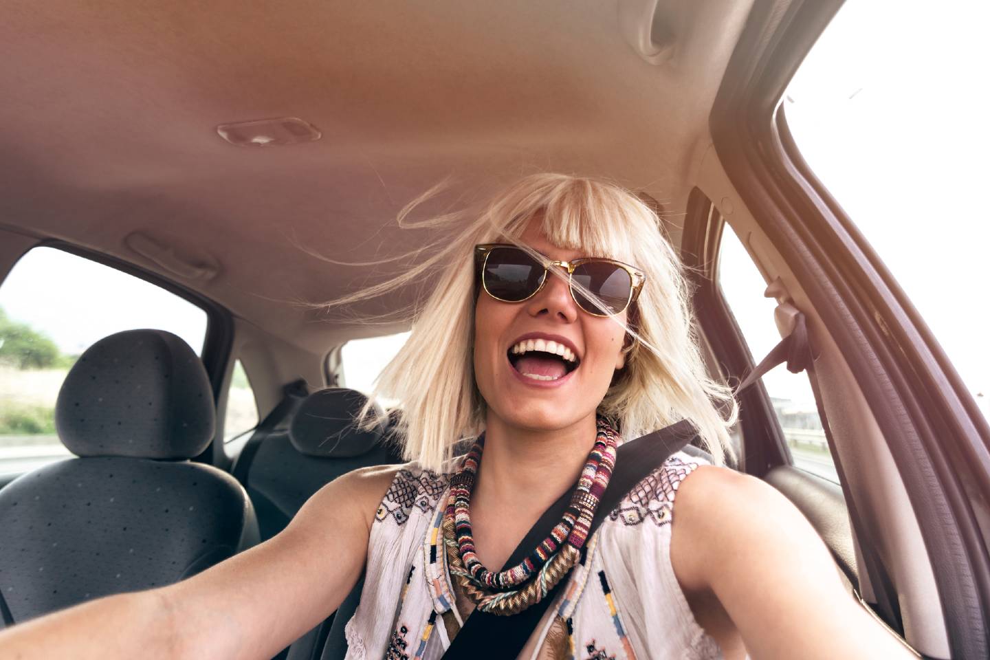 Eine Dame mit blondem Pagenschnitt sitzt am Steuer ihres Autos und sieht sehr fröhlich aus. 