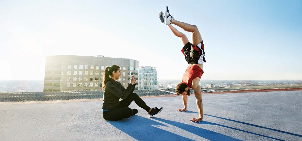 Auf dem Dach eines Hochhauses macht ein sportlicher Mann einen Handstand, eine junge Frau sieht ihm zu.