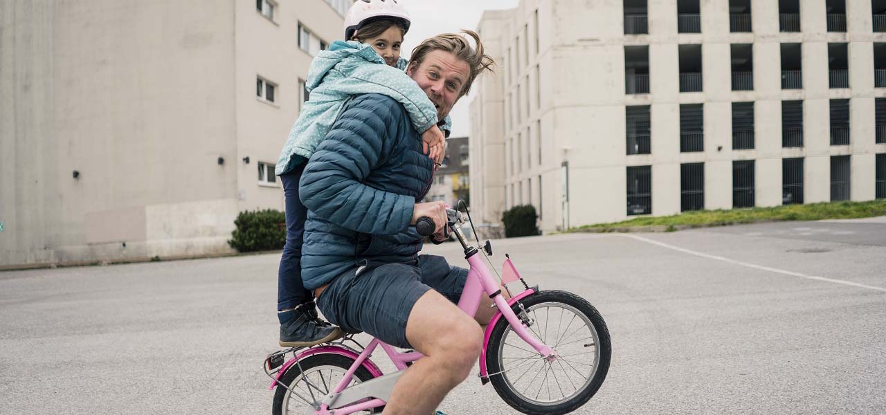 Vater sitzt auf einem Kinderfahrrad, die kleine Tochter steht auf dem Sattel und hält sich an ihm fest.