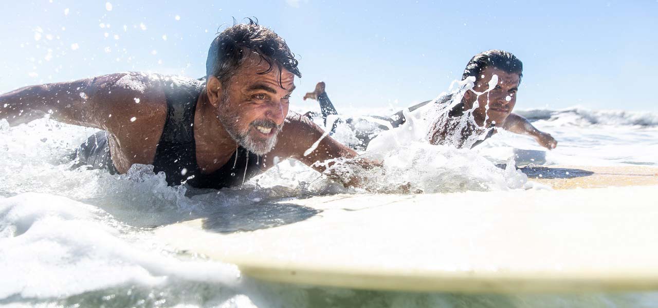 Zwei sportliche Männer mittleren Alters liegen auf Surfbrettern und paddeln im Meer. 