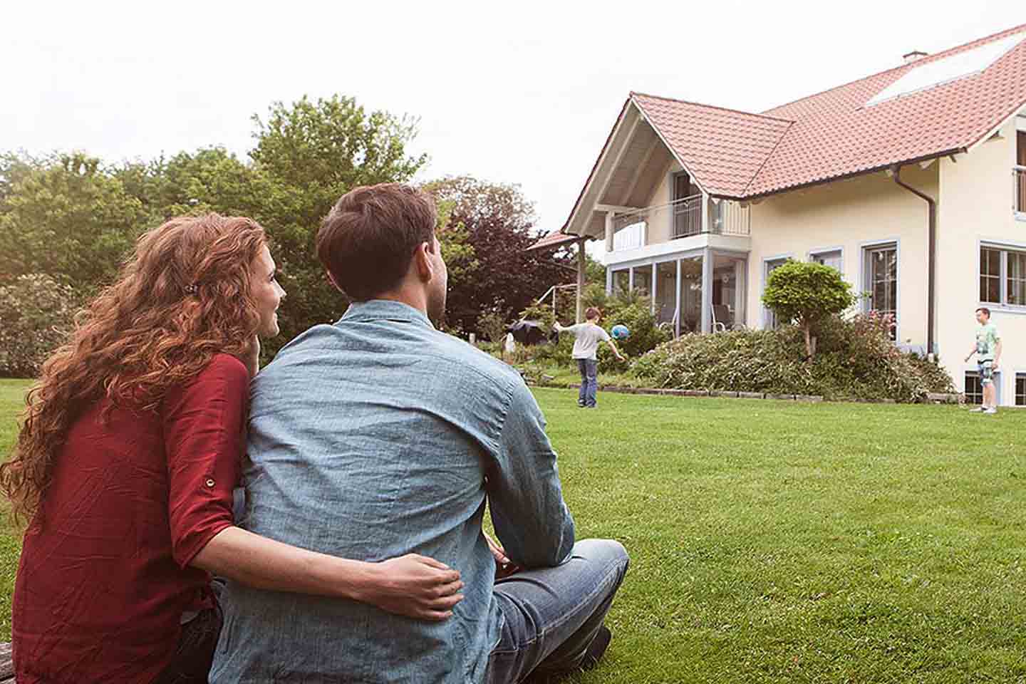 Ein Paar sitzt im Garten mit Blick auf ein Haus und sieht den Kindern beim Spielen zu.