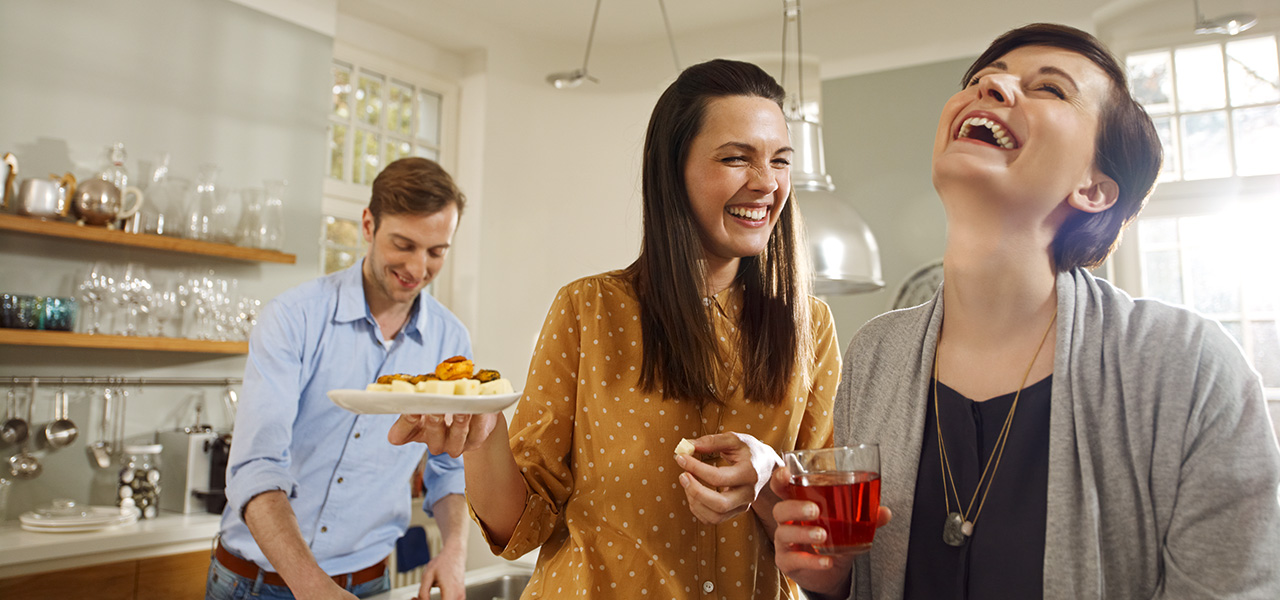 In einer Küche richtet ein Mann Essen während die Frauen miteinander lachen. Sie halten ein Getränk und Snacks.