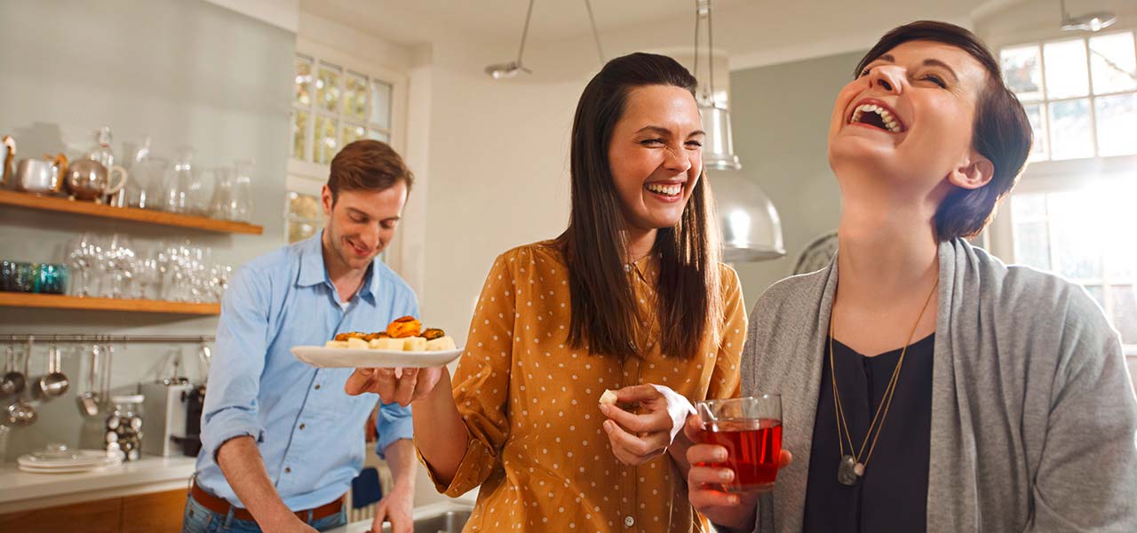 In einer Küche richtet ein Mann Essen während die Frauen miteinander lachen. Sie halten ein Getränk und Snacks.