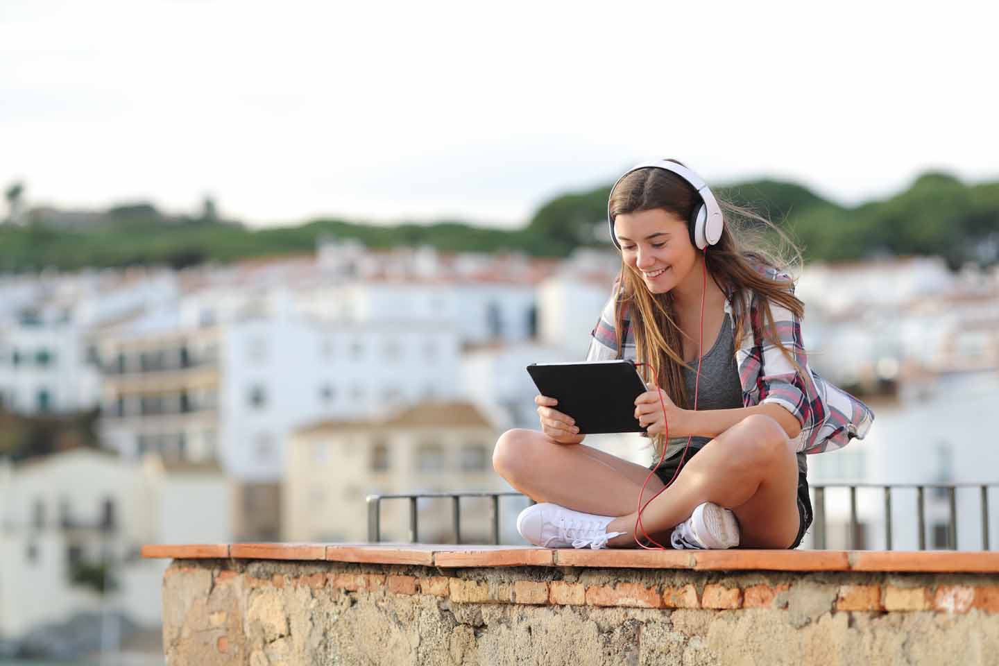 Eine junge Frau sitzt mit Kopfhörern und Tablet auf einem Dach und videotelefoniert.
