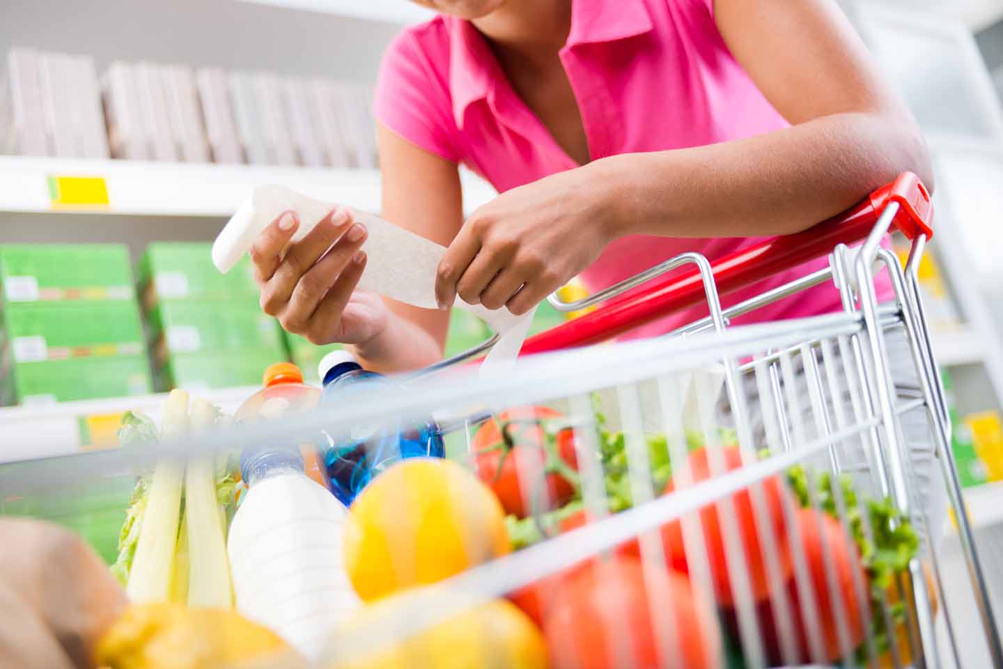 Eine Frau prüft einen Kassenbon. Dazu lehnt sie sich über ihren Einkaufswagen mit frischem Obst und Wasser.