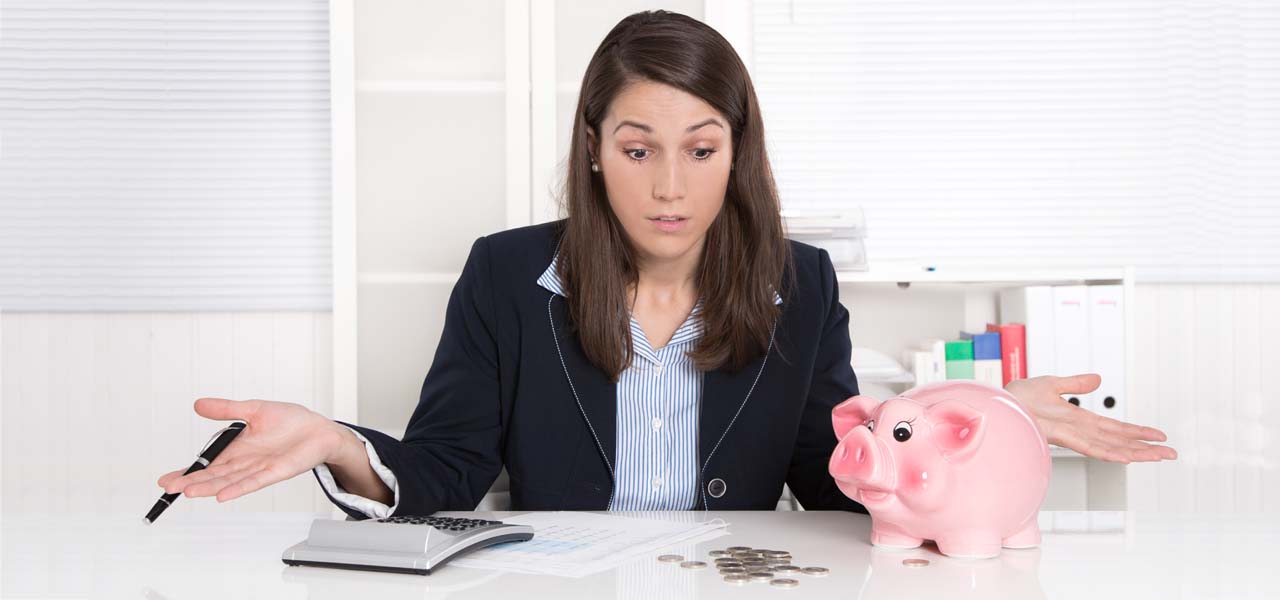 Eine Frau rechnet am Taschenrechner etwas nach und ist entsetzt. Ein Sparschwein und Münzen liegen auf dem Tisch.