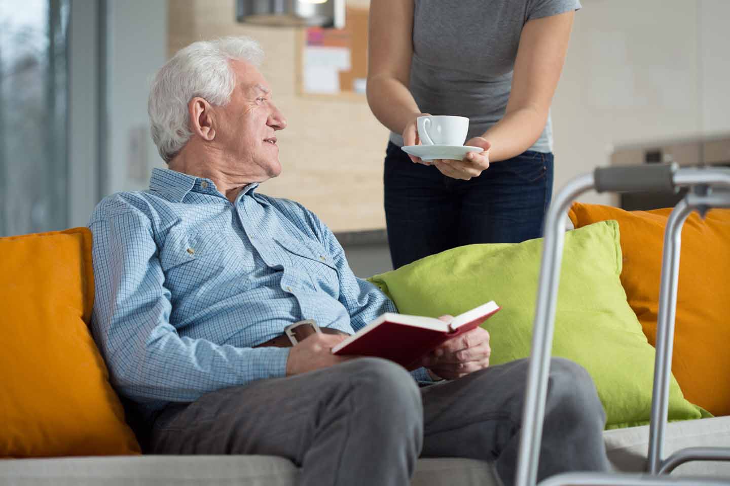 Ein Mann mit Gehhilfe bekommt eine Tasse Tee gereicht während er ein Buch liest.