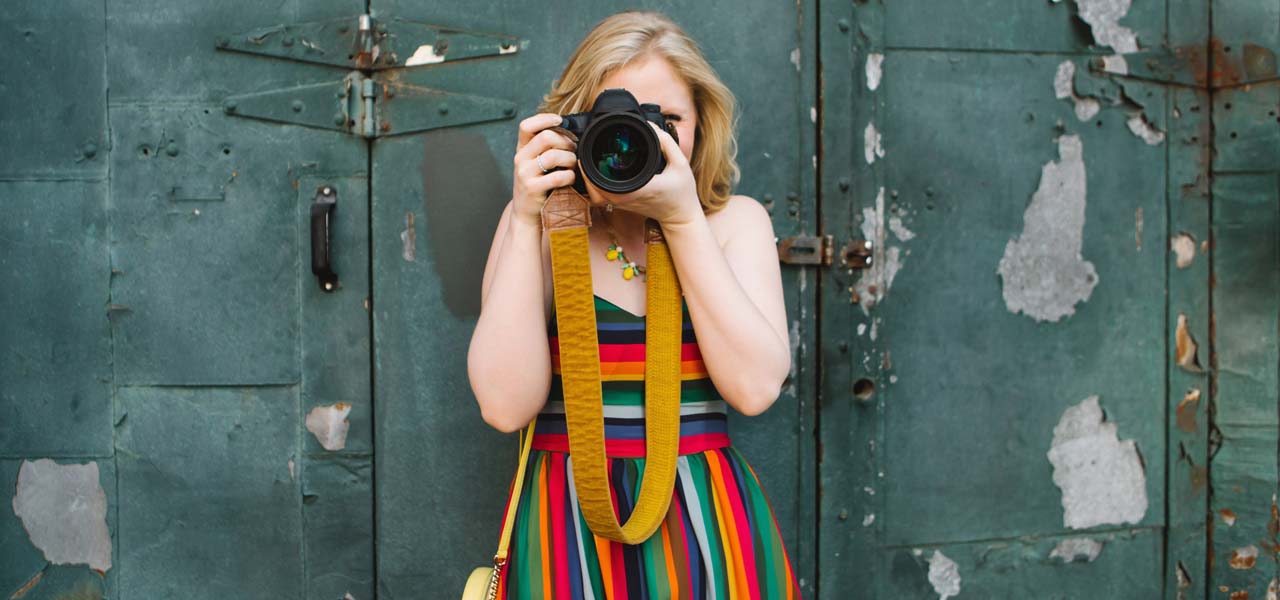 Eine Frau im bunten Kleid steht vor Metalltüren, von denen Farbe abplatzt und macht Fotos mit einer Spiegelreflexkamera.