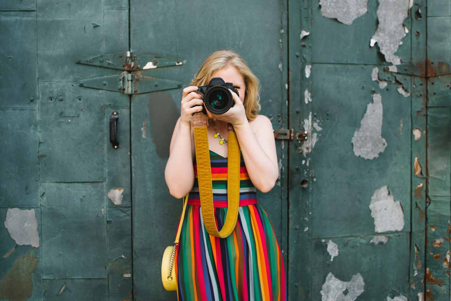 Eine Frau im bunten Kleid steht vor Metalltüren, von denen Farbe abplatzt und macht Fotos mit einer Spiegelreflexkamera.