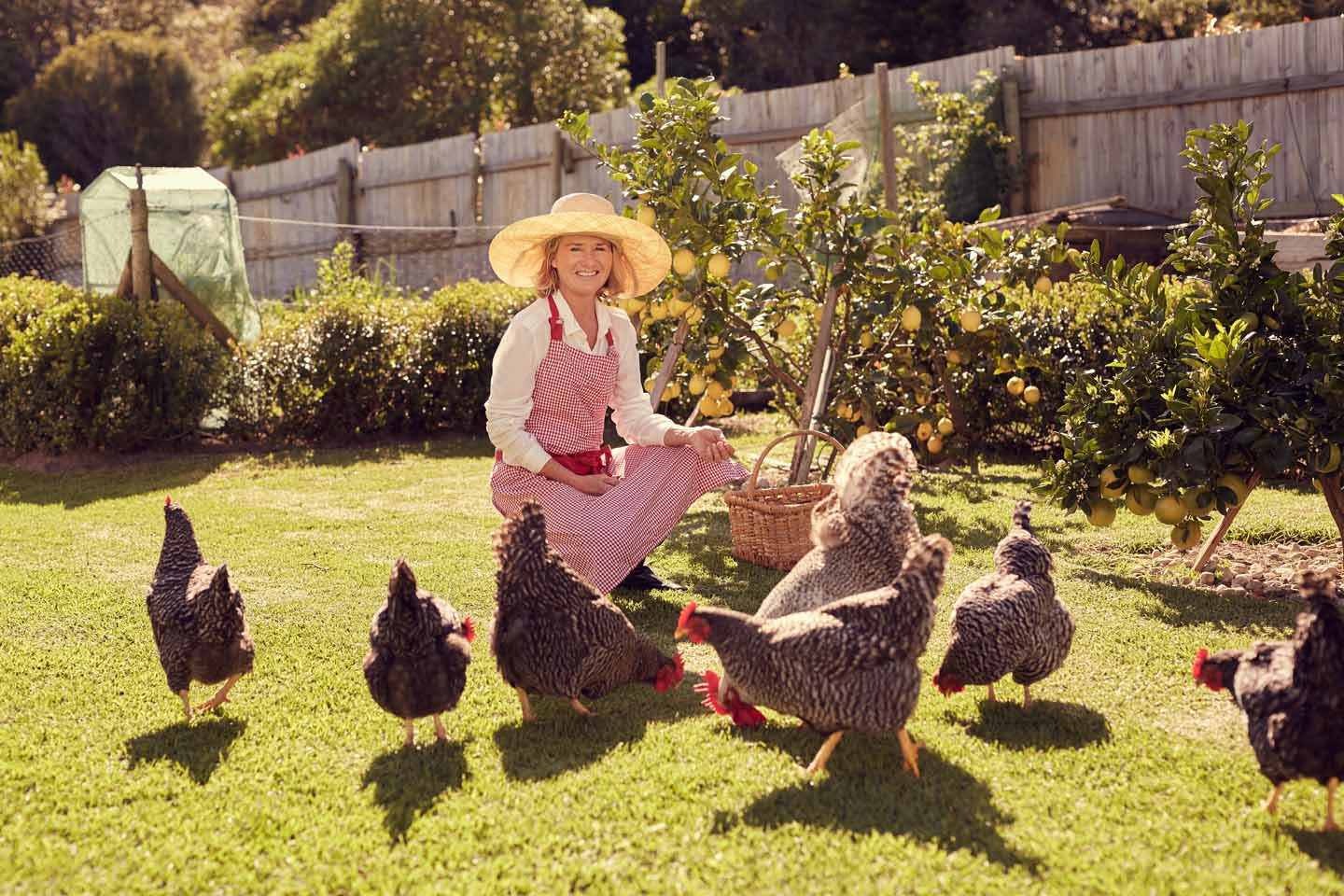 Eine Frau mit Hut füttert fröhlich die Hühner in ihrem Garten.