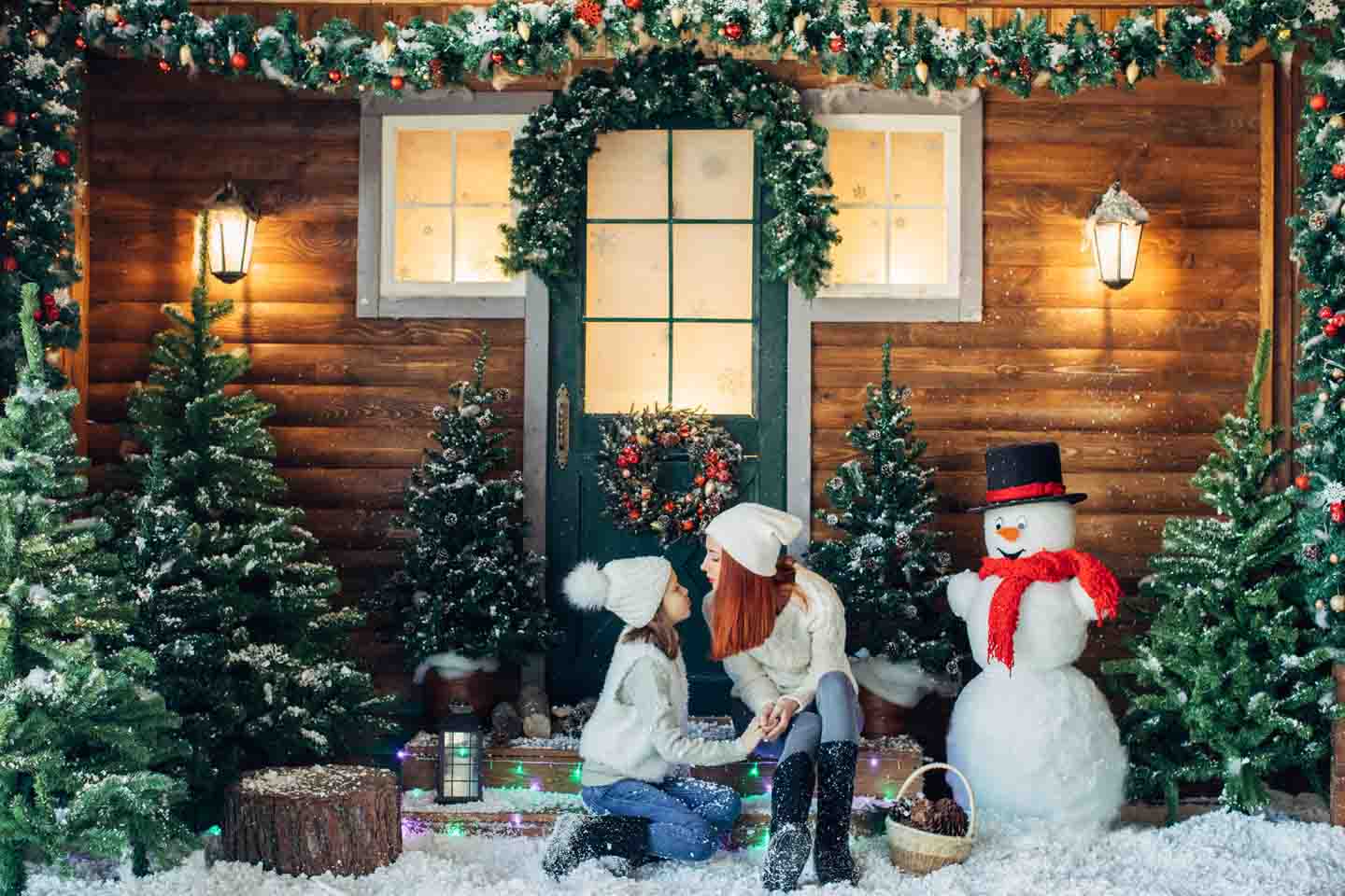 Mutter und Tochter sitzen auf der Treppe vor einem Haus, es schneit, ein Schneemann und Tannenbäume stehen herum.