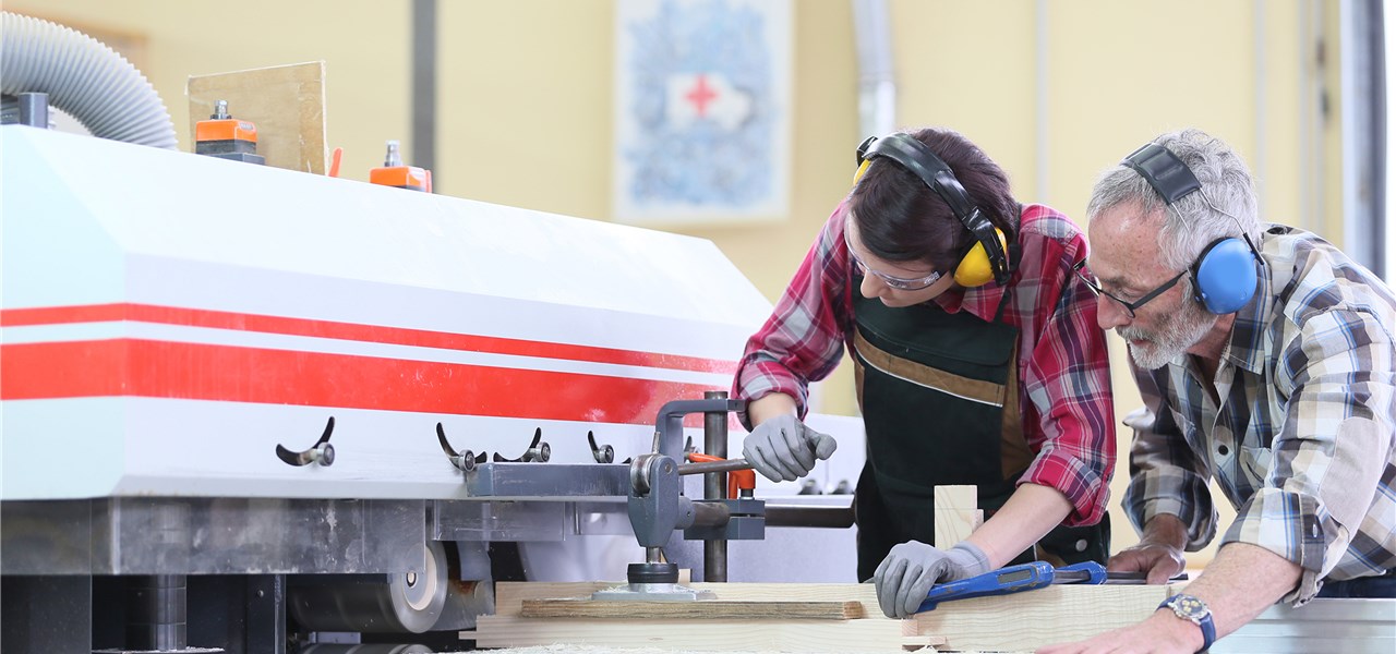 Mann und Jugendlicher in Schutzkleidung arbeiten an einer Maschine zur Holzverarbeitung.