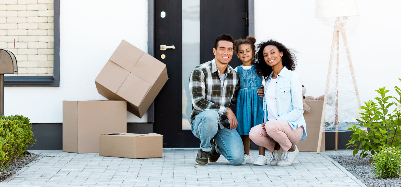 Junge Familie Arm in Arm vor einer Haustür, neben der Kartons stehen.