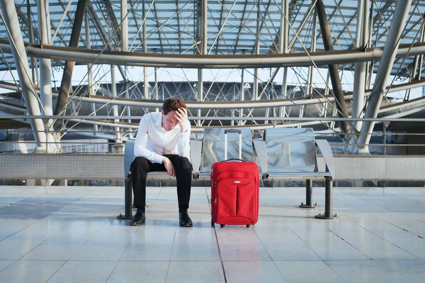 Ein Mann sitzt wartend auf einer Bank und stützt seinen Kopf auf seiner Hand auf. Neben ihm steht ein roter Koffer.