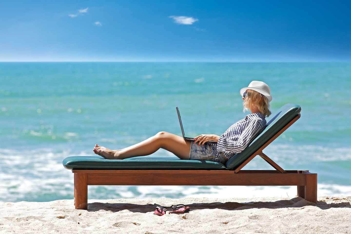 Eine Frau auf einer Liege am Strand mit Laptop auf dem Schoß.