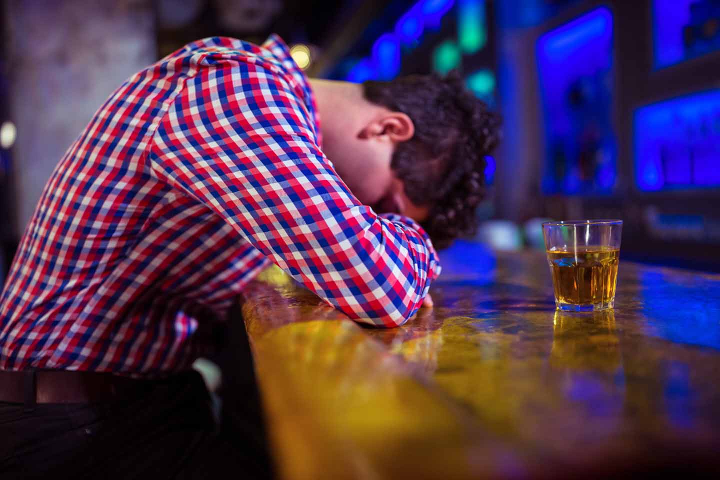 Ein Mann mit Karohemd liegt mit dem Kopf auf einem Tresen und schläft. Neben ihm steht ein Drink. 