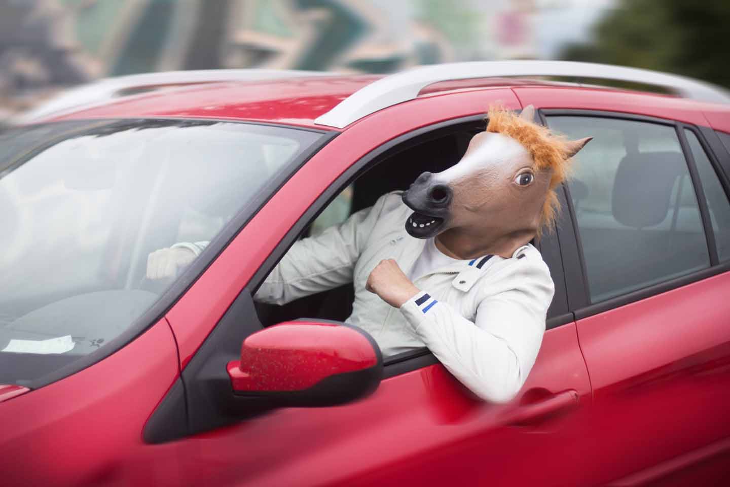 Aus dem Fenster eines roten Autos lehnt sich ein Rennfahrer mit Pferdemaske auf dem Kopf.