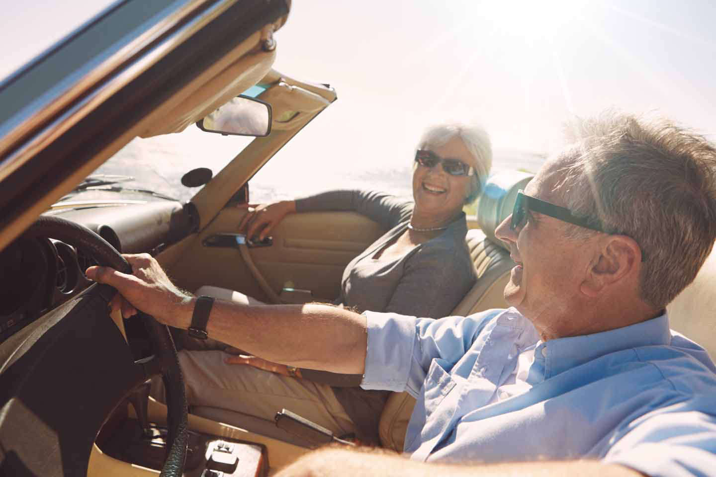 Ein älteres Paar sitzt im Cabrio. Beide tragen Sonnenbrillen und lachen.