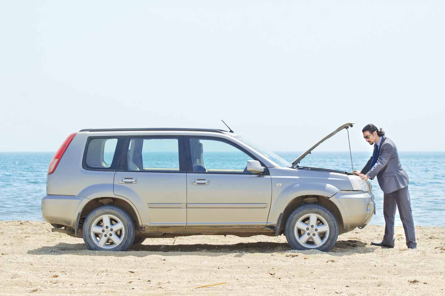 Am Sandstrand steht ein Mann im Anzug mit Sonnenbrille an der offenen Motorhaube seines Wagens. 
