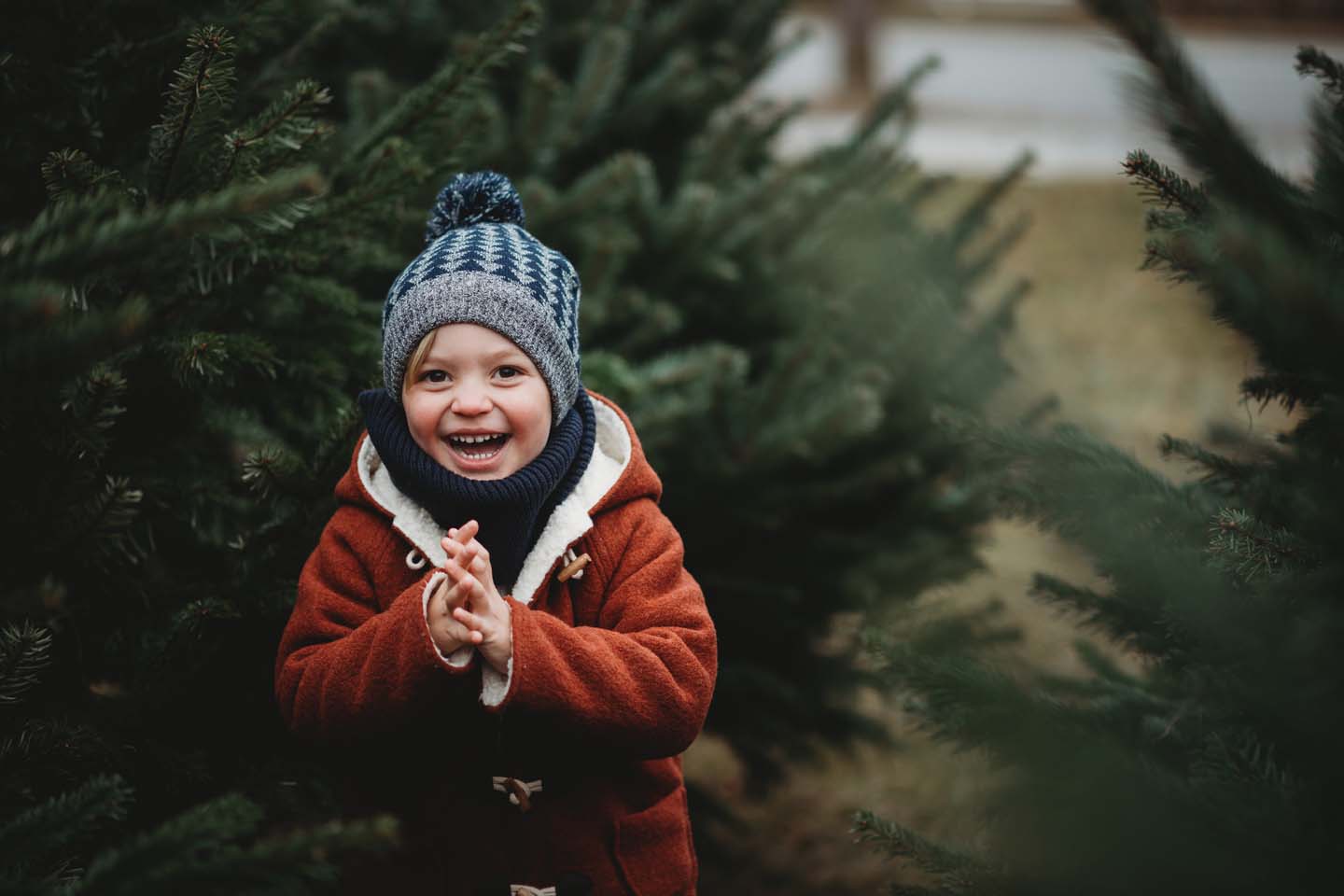 Ein lachendes kleines Kind mit dicker Winterkleidung steht vor vielen Tannenbäumen.