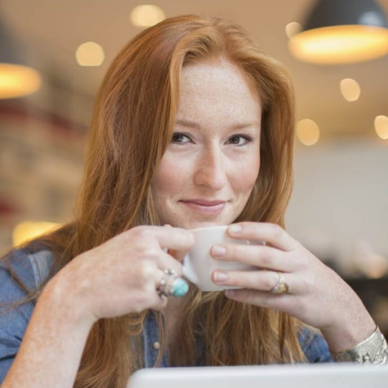 Junge Frau sitzt lächelnd mit Tasse in beiden Händen am Laptop.