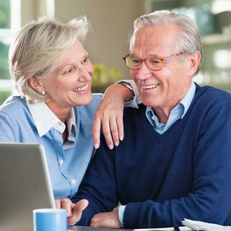 Älterer Mann und ältere Dame sitzen vertraut mit Laptop am Küchentisch und lächeln.