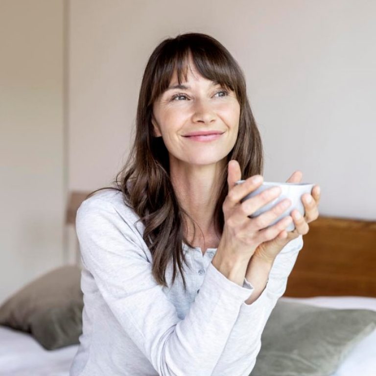 Entspannte Frau sitzt mit einer Tasse Kaffee auf dem Bett zu Hause.