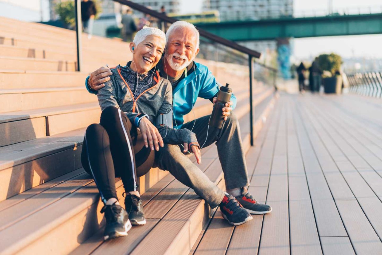Ein älteres Paar sitzt in Sportkleidung auf breiten Holzstufen. Beide haben Kopfhörer in den Ohren und der Mann hält eine Trinkflasche in der Hand.