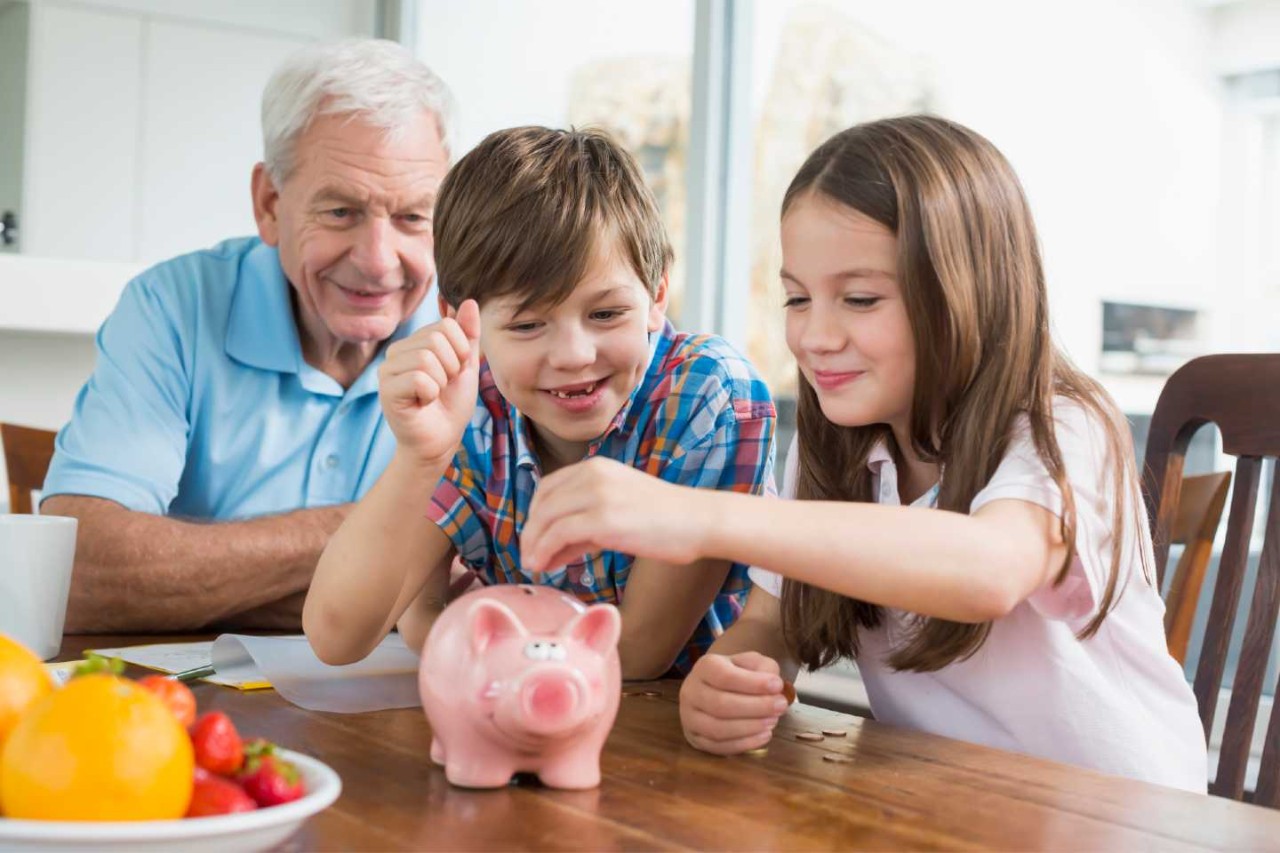 Ein kleiner Junge und ein kleines Mädchen sitzen mit dem Opa am Küchentisch und füttern ein Sparschwein mit Münzen.
